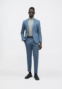 Világoskék, testhezálló szabású öltöny, szürke garbó fölött viselve. Fekete cipővel párosítva, tiszta, modern megjelenést kölcsönözve.