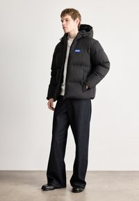 Μαύρο μπουφάν με κουκούλα, με μπροστινό φερμουάρ και λογότυπο. Συνδυασμένο με φαρδιά σκούρα τζην παντελόνια και μαύρα παπούτσια.