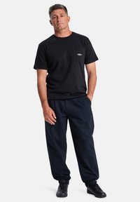 Zwarte katoenen t-shirt met korte mouwen en een borstzak, gecombineerd met loszittende donkerblauwe broek. Model draagt zwarte sneakers.
