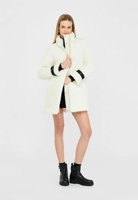 Witte jas van namaakbont met een hoge kraag, zwarte manchetten en twee grote voorzakken, gecombineerd met zwarte enkellaarsjes.