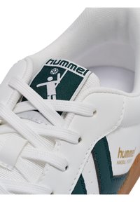 Hvid sneaker med grønne accenter og logo, med en tekstureret læderoverdel, flade snørebånd og en gummisål. Bemærkelsesværdig håndboldgrafik på tungen.