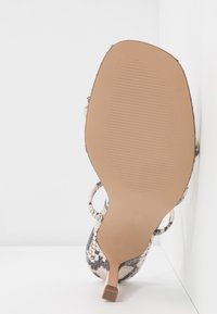 Semelle marron d'une sandale à talon haut avec un motif de bride en peau de serpent, présentée par le dessous sur fond blanc.