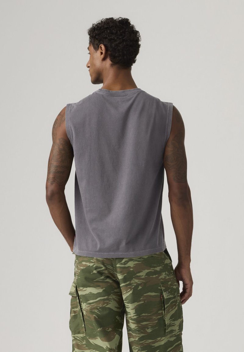 Homme debout, dos à la caméra, portant un débardeur gris et un short cargo camouflage vert, tatouages visibles sur les bras.