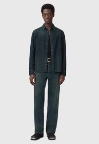 Camicia in denim verde scuro e jeans coordinati, entrambi con una finitura leggermente sbiadita. Cintura nera con fibbia in argento, scarpe nere lucide.