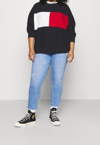 Marineblauer Pullover mit roten und weißen geometrischen Streifen, kombiniert mit hellblauen, abgeriebenen Jeans und schwarzen High-Top-Sneakern.