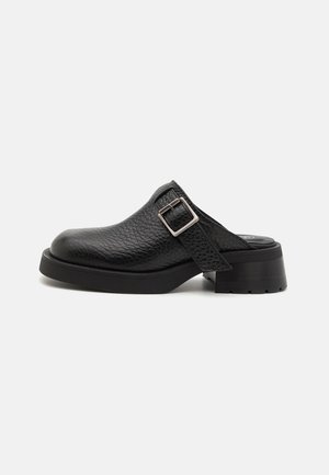 Sabots en cuir noir avec une finition texturée, bride à boucle réglable et un talon épais. Design de la pointe lisse et arrondie avec une semelle noire solide.