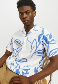 Camisa blanca de manga corta con patrones florales en azul y beige, material ligero, con botones en el frente y un corte holgado, combinada con pantalones beige.