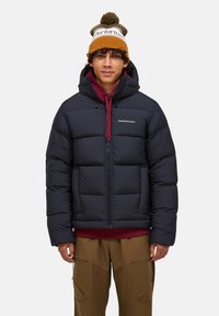 Sort polstret jakke med lynlås, der har en hætte og store quiltede sektioner. Bæres over en bordeaux hoodie. Brun strikket beanie med pompon.