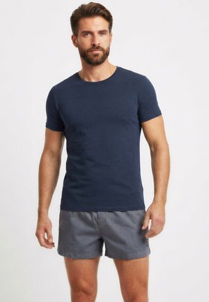 Mann mit Bart, der ein dunkelblaues T-Shirt und graue Shorts trägt, steht vor einem schlichten weißen Hintergrund.