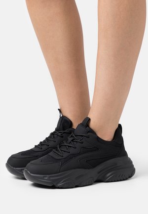 Ben iført sorte chunky sneakers med tykke såler, mesh- og læderpaneler samt snørebånd, vist mod en ensartet lys baggrund.