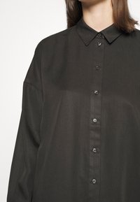 Dark button-up shirt made of smooth fabric. Features a pointed collar, long sleeves, and round buttons down the front.