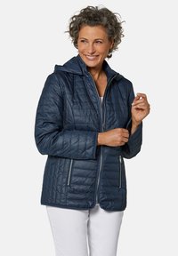 Veste matelassée bleu marine avec capuche, dotée de deux poches latérales zippées, d'une texture lisse et d'une coupe ajustée.