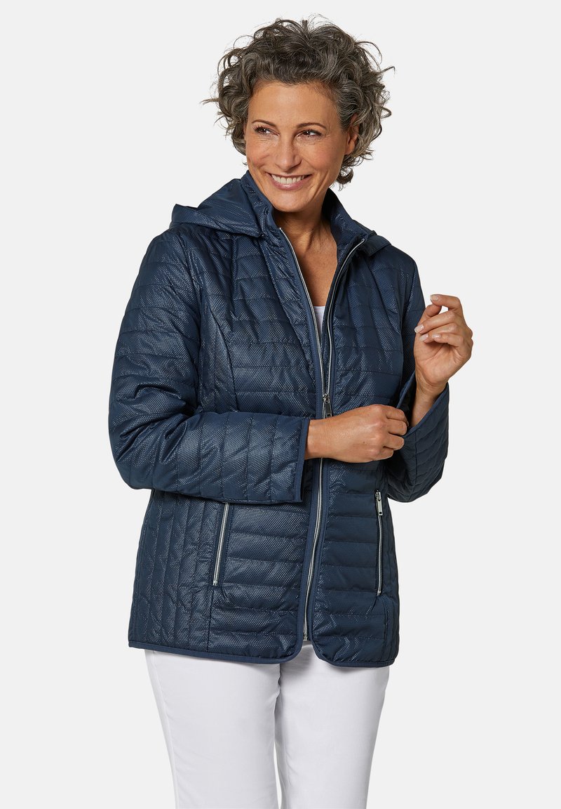 Veste matelassée bleu marine avec capuche, dotée de deux poches latérales zippées, d'une texture lisse et d'une coupe ajustée.