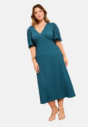 Kvinde iført en aqua tekstureret midikjole med flagrende ærmer og V-udskæring, kombineret med beige strop-sandaler og en guldbøjle.