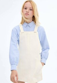 Crème kleurige denim salopette-jurk met twee voorzakken, gecombineerd met een blauw-wit gestreept shirt met lange mouwen. Eenvoudig ontwerp.