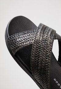 Svart halksandaler med flätad metallrem och en texturerad fotbädd. Platt sula, minimalistisk design och släta detaljer.