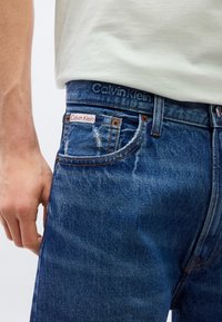 Μπλε τζιν παντελόνι Calvin Klein με ελαφριά ξεθώριασμα, σχεδίαση με πέντε τσέπες και κόκκινη ετικέτα λογότυπου στην μπροστινή τσέπη. Η ζώνη έχει ανάγλυφο λογότυπο.