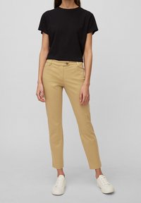 Pantalons en coton beige à coupe slim, avec poches latérales et fermeture à bouton, associés à un t-shirt noir à manches courtes et des baskets blanches.