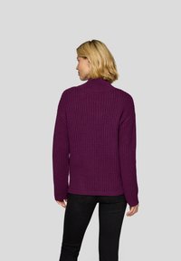 Lila gestrickter Pullover mit hohem Kragen, lockerer Passform und gerippter Textur, kombiniert mit schwarzen Hosen. Die Rückansicht zeigt fallende Schultern.