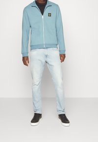 Chaqueta azul claro con cremallera y cuello alto, con un logotipo en la parte frontal, combinada con jeans de mezclilla claros y zapatillas negras. Tejido suave y ajuste casual.