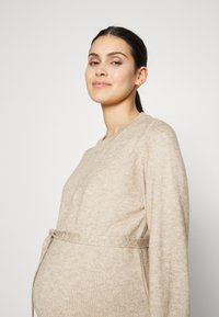 Beige neulepaita odottaville äideille, jossa on pitkät hihat, pyöreä kaula-aukko ja solmittava vyötäröyksityiskohta. Pehmeä tekstuuri ja rento istuvuus.