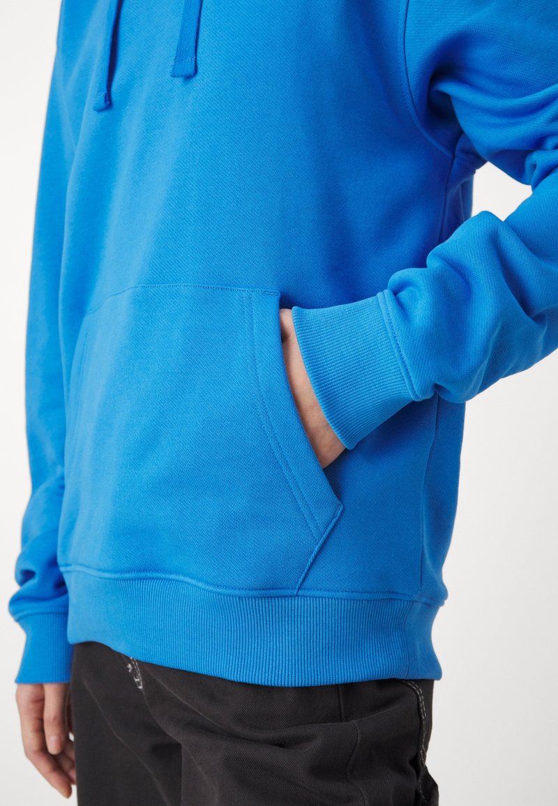 Personne portant un sweat à capuche bleu vif avec une main dans la poche avant, associé à un pantalon noir.
