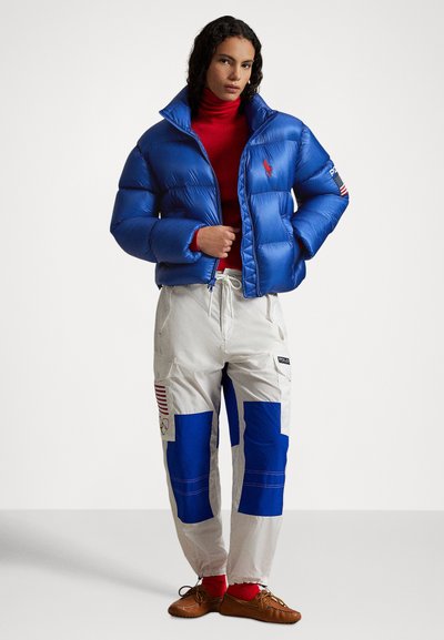 Blue puffer jacket with a raised collar and red polo logo, paired with white and blue cargo pants featuring Olympic patches and pockets.
