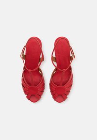 Paire de sandales rouges en cuir à bout ouvert avec un design circulaire à l'avant, des brides à la cheville et des fermetures à boucle dorée sur fond blanc.