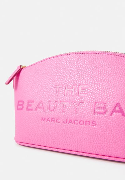 Vaaleanpunainen nahkainen kosmetiikkalaukku, jossa on kaareva yläosa, kohokuvioitu teksti "THE BEAUTY BAG" ja "MARC JACOBS", kultainen vetoketju ja vetotanko.