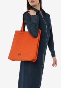 Borsa a tracolla arancione a costine con spalline larghe e forma rettangolare. Presenta un piccolo logo nero sul fronte ed è realizzata in materiale testurizzato.