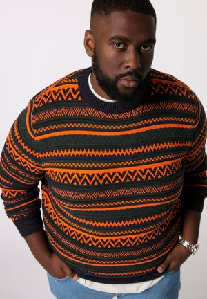 Gebreid sweater met een multikleurig geometrisch patroon in oranje, groen en marineblauw. Normale halslijn met geribbelde manchetten, gedragen over een wit shirt.