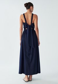 Kvinna i en marinblå ärmlös maxiklänning med volangband och en stor rosett knuten vid den öppna ryggen, i kombination med platta sandaler.
