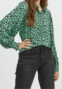 Blusa verde con un motivo astratto nero, dotata di un colletto e maniche lunghe a sbuffo. Abbinata a jeans neri a vita alta.