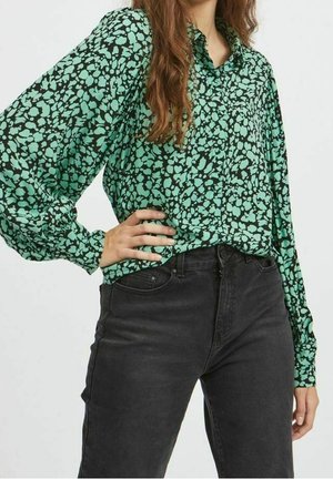 Blusa verde con un motivo astratto nero, dotata di un colletto e maniche lunghe a sbuffo. Abbinata a jeans neri a vita alta.