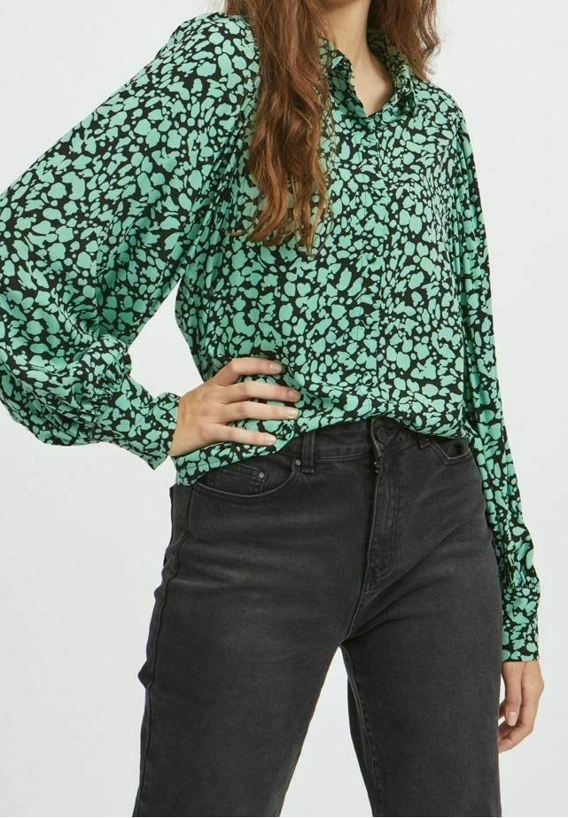 Blusa verde con un motivo astratto nero, dotata di un colletto e maniche lunghe a sbuffo. Abbinata a jeans neri a vita alta.