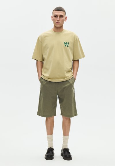 Jeune homme portant un t-shirt beige oversize avec le logo vert "W", un short vert olive, des chaussettes crème et des chaussures noires à lacets, debout les mains dans les poches.