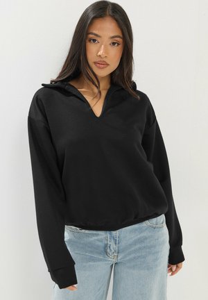 Ung kvinde med langt mørkt hår iført en sort V-hals trøje og lyseblå jeans, stående mod en ensfarvet lys baggrund.