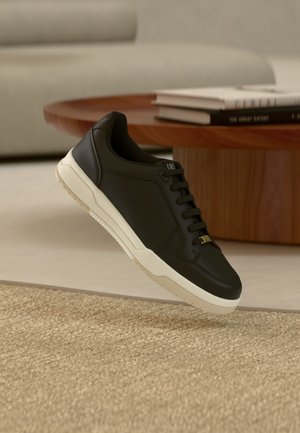 Baskets basses noires avec semelle blanche flottant au-dessus d'un tapis beige texturé dans un salon moderne avec une table basse en bois.