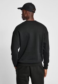 Man som bär svart keps, svart långärmad sweatshirt och svarta byxor, står med ryggen mot kameran framför en enkel bakgrund.