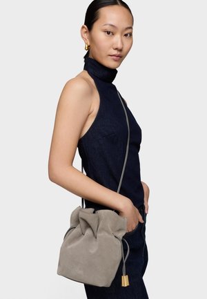 Femme aux cheveux lisses portant un haut sans manches sombre et tenant un sac bourse gris en daim avec des accents dorés sur son épaule.