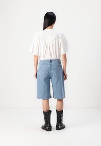 Blå denimshorts med høy midje, som slutter rett over kneet, sammen med en hvit bluse med volangdesign på ryggen og svarte støvler.