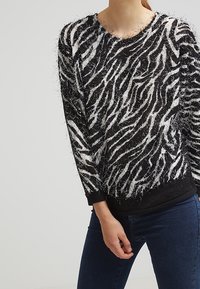 Svetr se zebrovaným vzorem v černobílé barvě, vyrobený z huňatého materiálu, s žebrovanými lemy na rukávech a spodním lemu. Design má kulatý výstřih a volný střih.