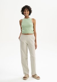 Hellgrünes, tailliertes, ärmelloses Oberteil; bequeme beige Hose; braune Rutschsandalen; Person steht vor einem weißen Hintergrund.