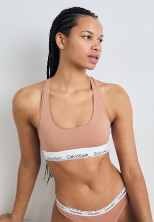 Sutien sport de culoare maro deschis, cu o bandă albă lată pe care este inscripționat logo-ul "Calvin Klein". Este un model ajustat, cu decolteu rotund și spate tip racer.