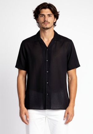 Uomo che indossa una camicia nera a maniche corte con colletto e pantaloni bianchi, in piedi davanti a uno sfondo semplice.