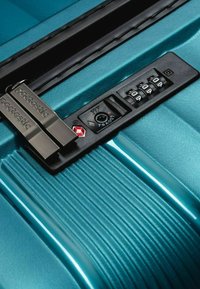 Maleta dura color verde azulado con un acabado texturizado, cerradura aprobada por la TSA y un marcador de combinación. Presenta acentos metálicos y áreas de diseño acanaladas.