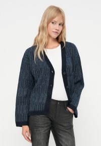 Tânără cu păr blond, îmbrăcată într-un cardigan în carouri bleumarin și blue, cămașă albă și pantaloni negri texturați, stând cu o mână în buzunar.