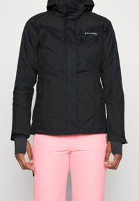 Svart isolerad jacka med huva, med en dragkedjeförsedd bröstficka, sidofickor och justerbara ärmar. Bärs med rosa byxor.