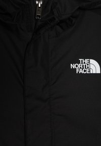 Svart, väderbeständig jacka med dragkedja som stängs, hög krage och en vit logotyp på vänster bröstområde. Slät tygerstruktur.