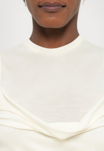 Cream-colored top with a smooth texture, featuring a round neckline and draped detailing at the front. Simple, form-fitting design.